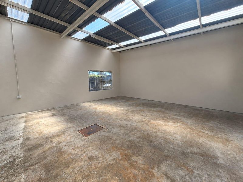 An empty, utilitarian commercial space featuring a concrete floor, exposed steel roof structure with translucent panels, and a single window with security bars.