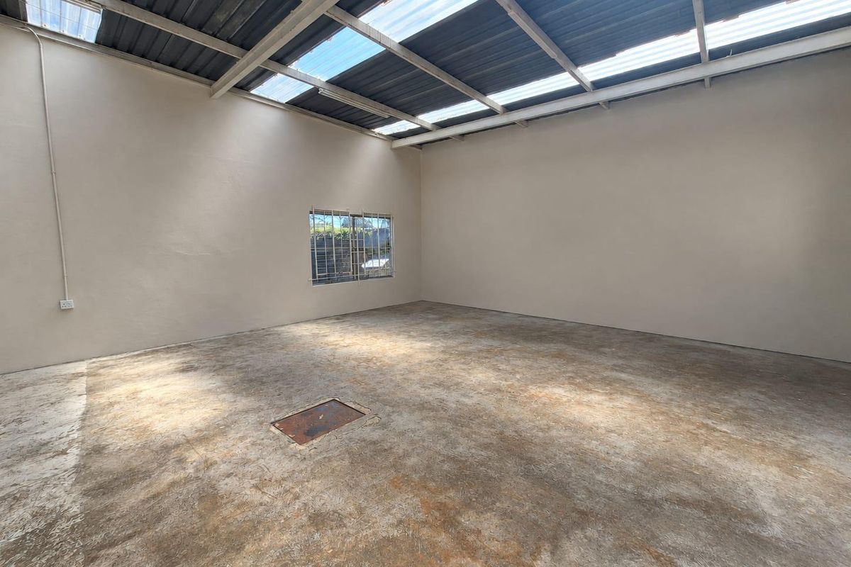 An empty, utilitarian commercial space featuring a concrete floor, exposed steel roof structure with translucent panels, and a single window with security bars.