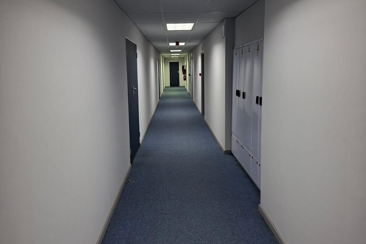 A long commercial office hallway featuring blue carpet flooring, suspended ceiling with recessed lighting, and a row of built-in storage lockers.