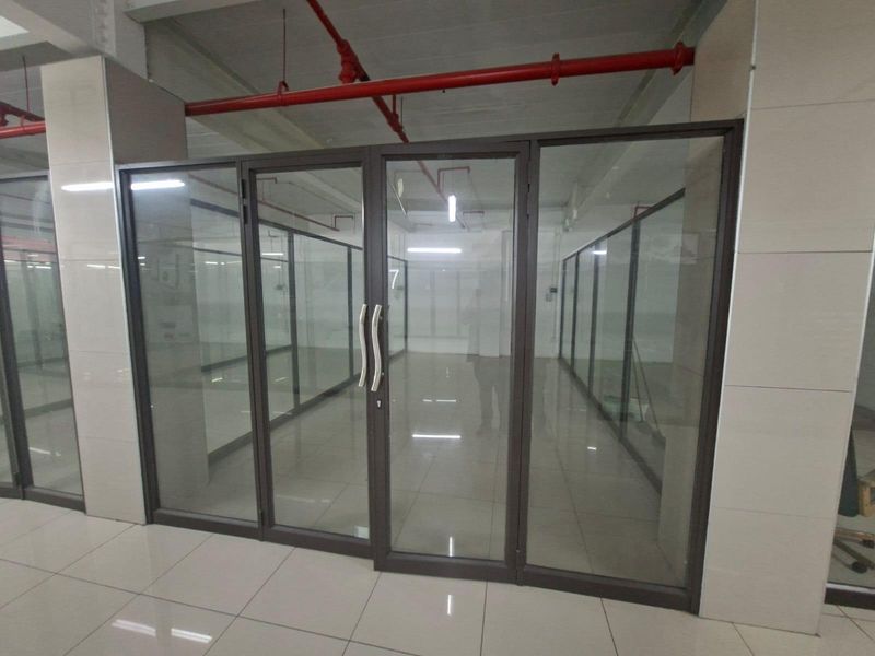 A modern, empty commercial office space featuring glass partition walls, tiled flooring, and exposed industrial-style fire sprinkler systems.