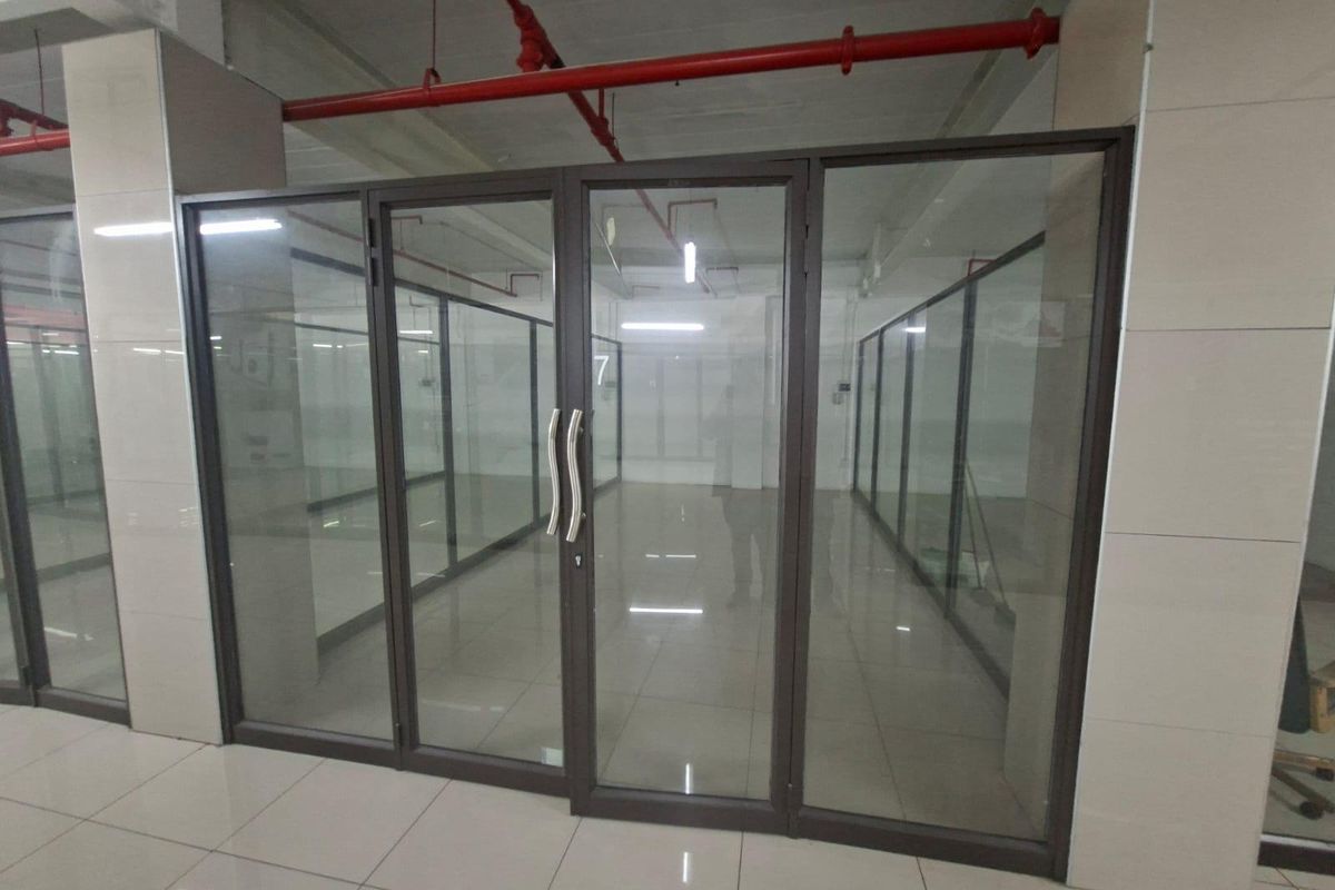 A modern, empty commercial office space featuring glass partition walls, tiled flooring, and exposed industrial-style fire sprinkler systems.