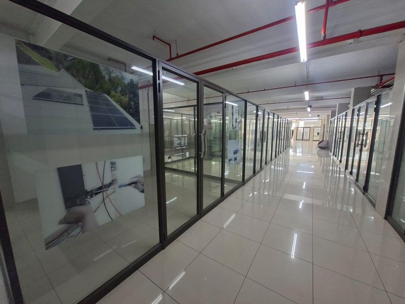 A modern commercial hallway featuring extensive glass partitioning, tiled flooring, and exposed industrial-style ceiling infrastructure with fire safety systems.