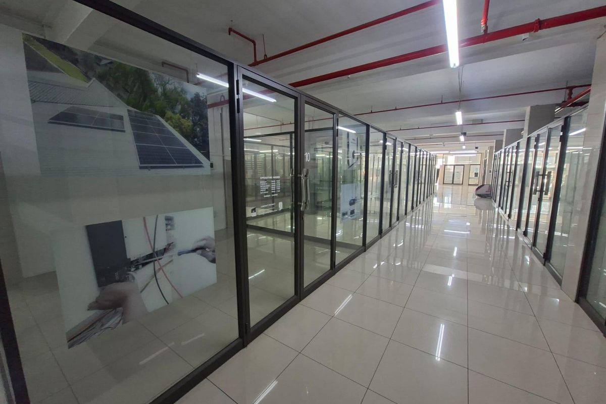 A modern commercial hallway featuring extensive glass partitioning, tiled flooring, and exposed industrial-style ceiling infrastructure with fire safety systems.