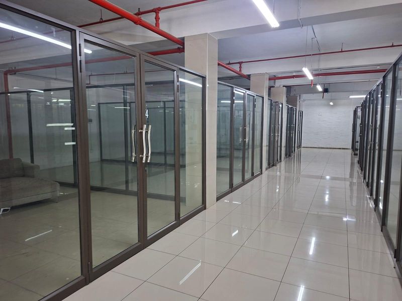 A commercial hallway featuring glass-partitioned office units, polished ceramic floor tiles, and exposed industrial-style fire sprinkler systems.