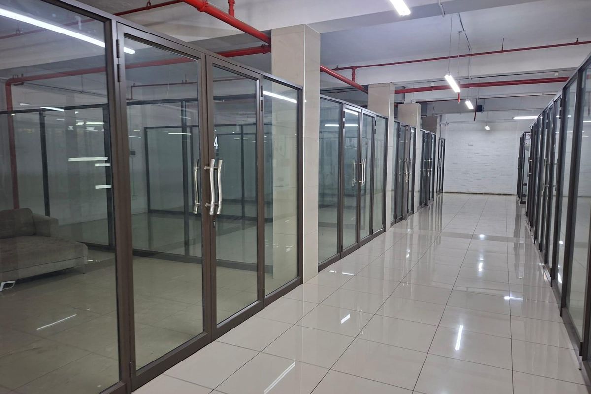 A commercial hallway featuring glass-partitioned office units, polished ceramic floor tiles, and exposed industrial-style fire sprinkler systems.