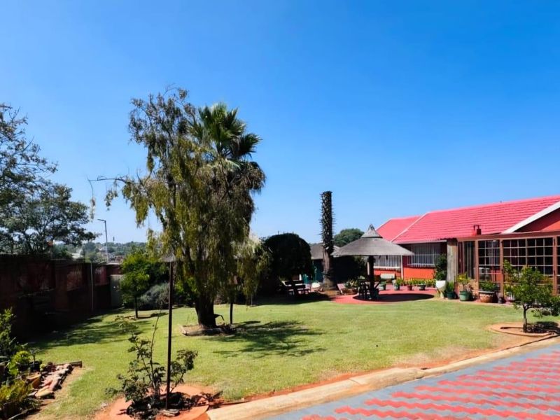 Spacious rear garden featuring a well-maintained lawn, paved driveway, and an enclosed patio area attached to the main house with a red metal roof.