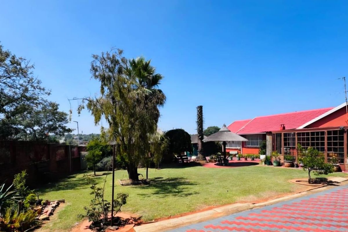 Spacious rear garden featuring a well-maintained lawn, paved driveway, and an enclosed patio area attached to the main house with a red metal roof.