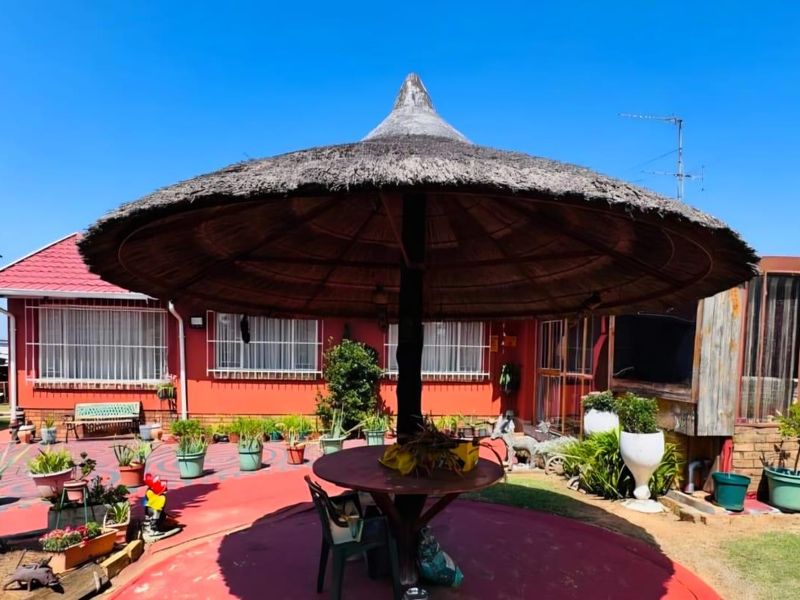 Rear exterior view featuring a traditional thatched lapa, paved patio area, and red-painted building facade with security bars on windows.