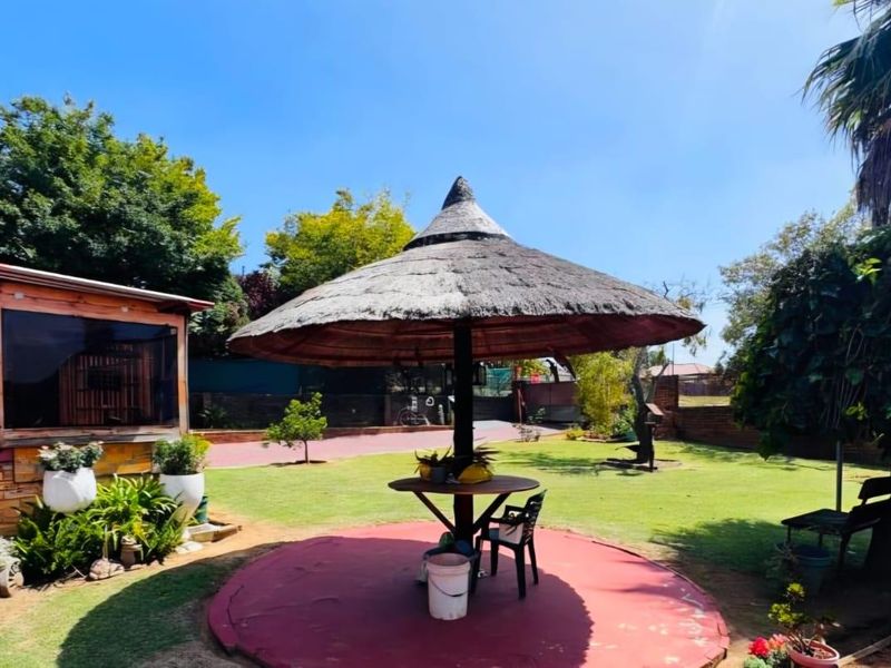 A spacious garden featuring a central thatched lapa, a circular paved entertainment area, and established lawn, offering potential for outdoor living.