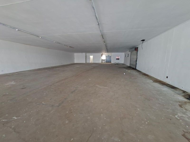 A large, open-plan commercial space featuring concrete flooring, fluorescent lighting, and a roller shutter door, suitable for various retail or industrial applications.