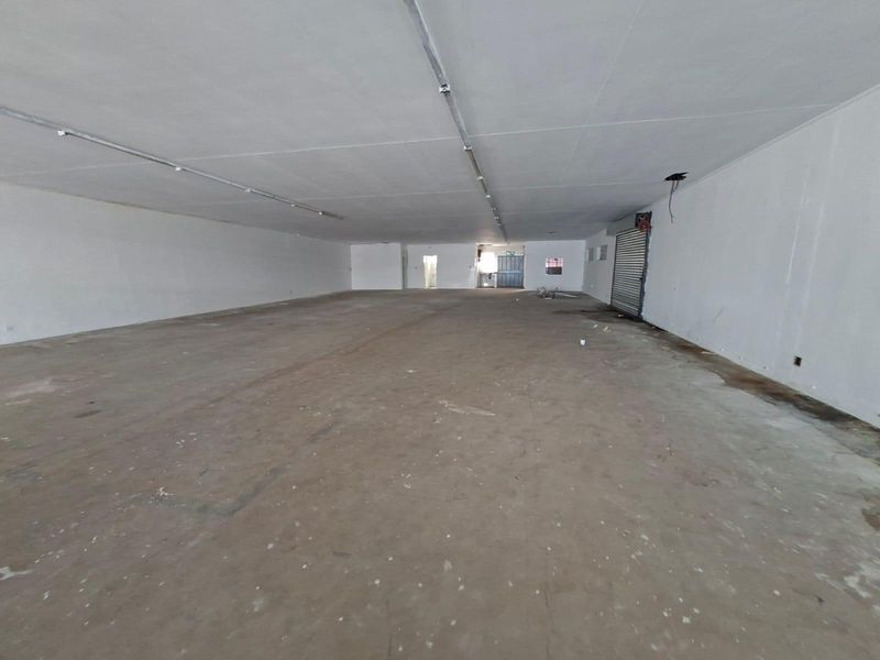 A large, vacant open-plan commercial space featuring concrete flooring, a roller shutter door, and basic overhead lighting, suitable for various retail or industrial uses.
