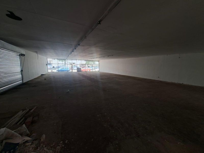 Spacious open-plan commercial unit featuring a large glass storefront, roller shutter door, and raw concrete flooring, suitable for various retail or office configurations.