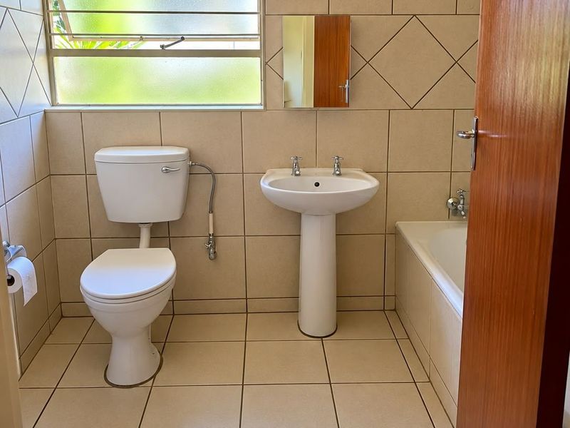 A functional bathroom featuring a pedestal basin, toilet, and bathtub with neutral wall and floor tiling and a louvered window for ventilation.
