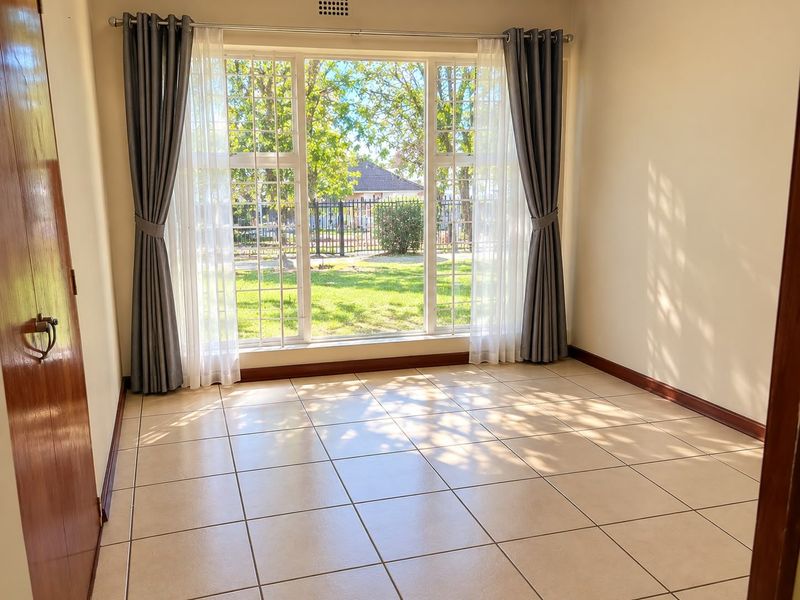 A bright and spacious empty living area featuring neutral tiled flooring, large windows with garden views, and classic wooden door finishes.