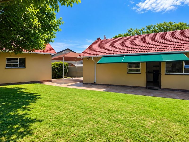 Rear view of the property featuring a well-maintained lawn, paved patio area, and a retractable awning over the main building entrance.