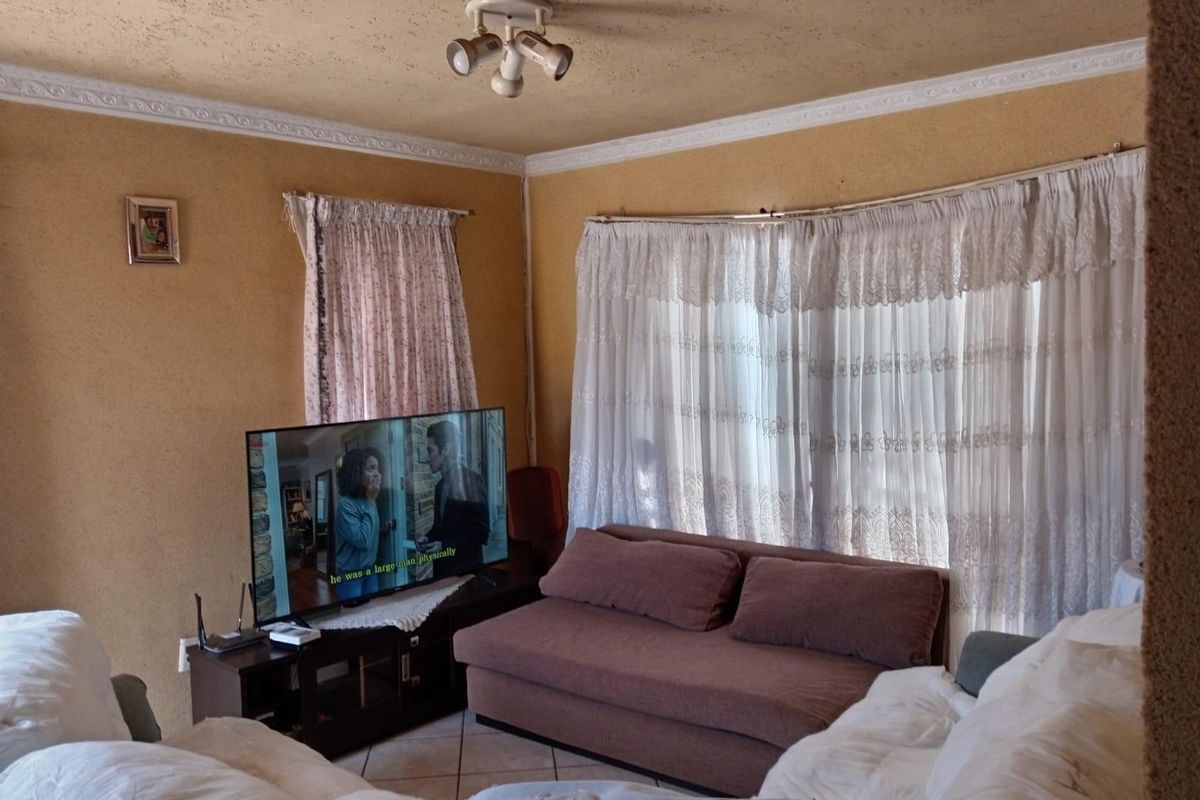 A living room featuring tiled flooring, decorative ceiling cornices, and a central light fixture, currently showing signs of wear on the wall finishes.