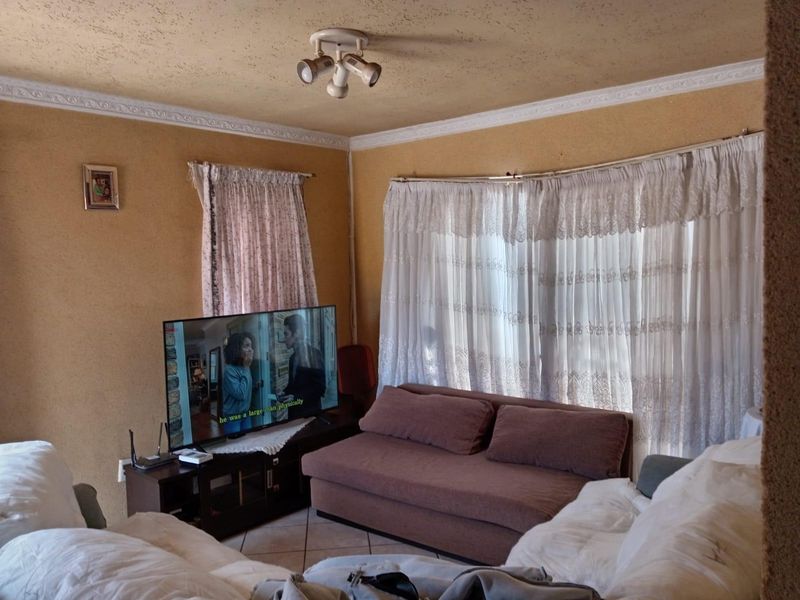 A living room featuring tiled flooring, decorative ceiling cornices, and a central light fixture, currently showing signs of wear on the wall finishes.