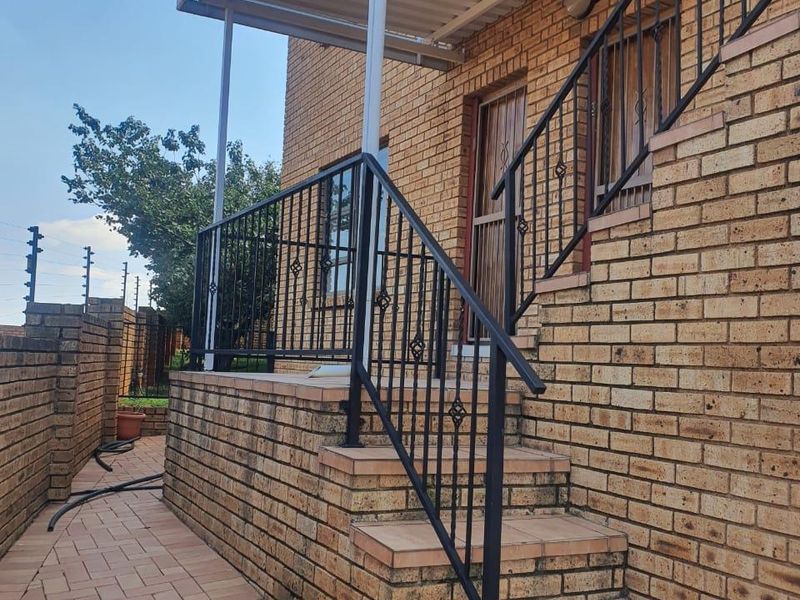 Rear exterior view of the apartment featuring a brick facade, a covered patio area with tiled steps, and secure metal railings.