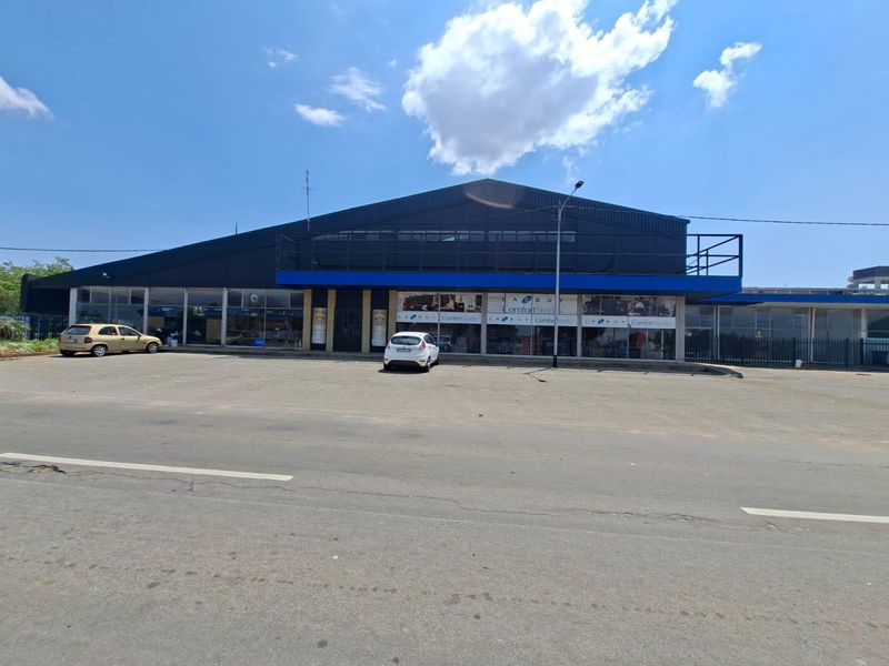 Front exterior view of a commercial building featuring a large glass storefront, metal cladding, and an expansive asphalt parking area.