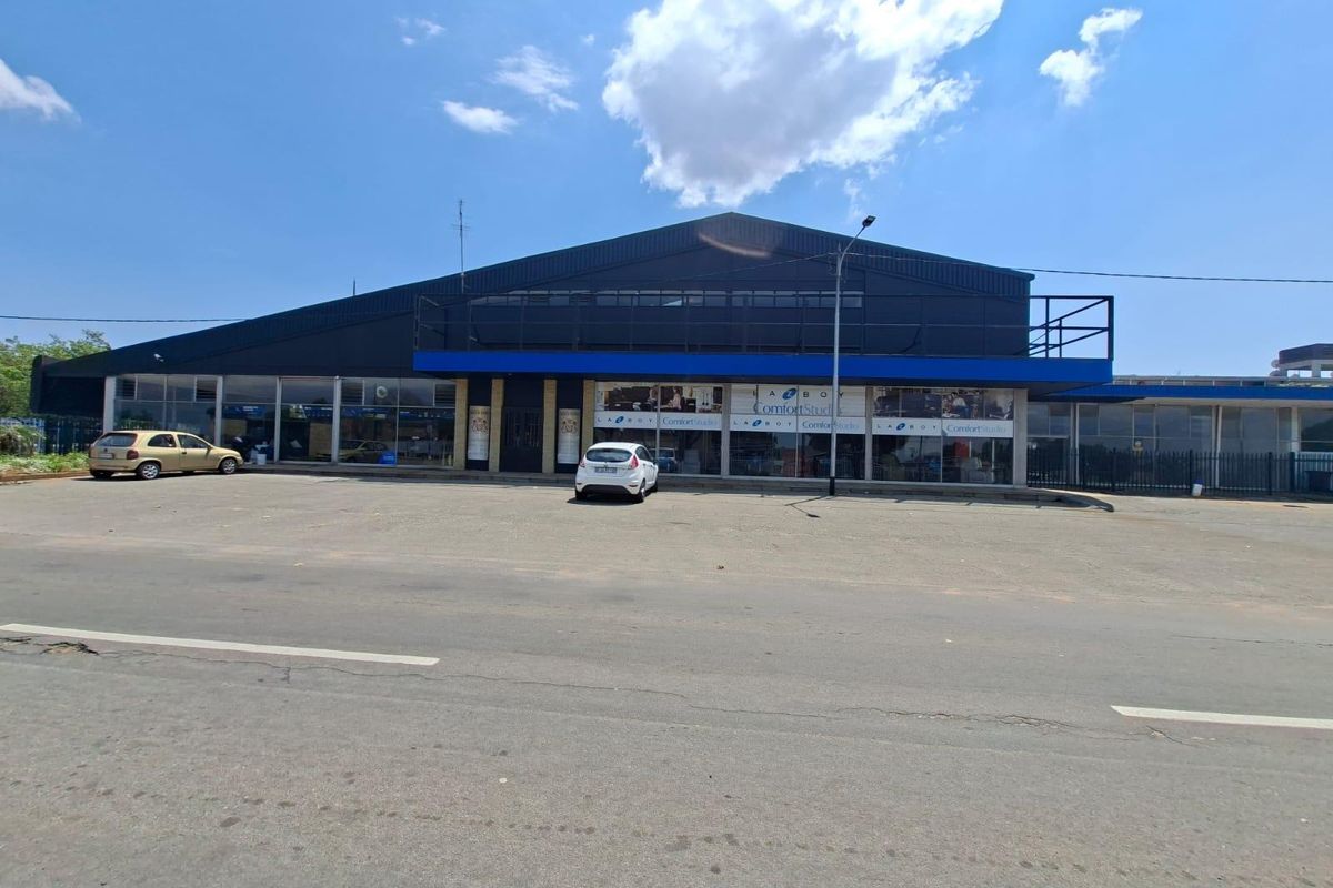 Front exterior view of a commercial building featuring a large glass storefront, metal cladding, and an expansive asphalt parking area.