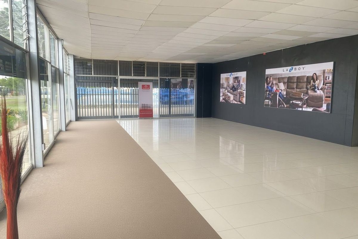 Spacious open-plan commercial showroom area featuring a mix of tiled and carpeted flooring, large glass windows, and a suspended ceiling.