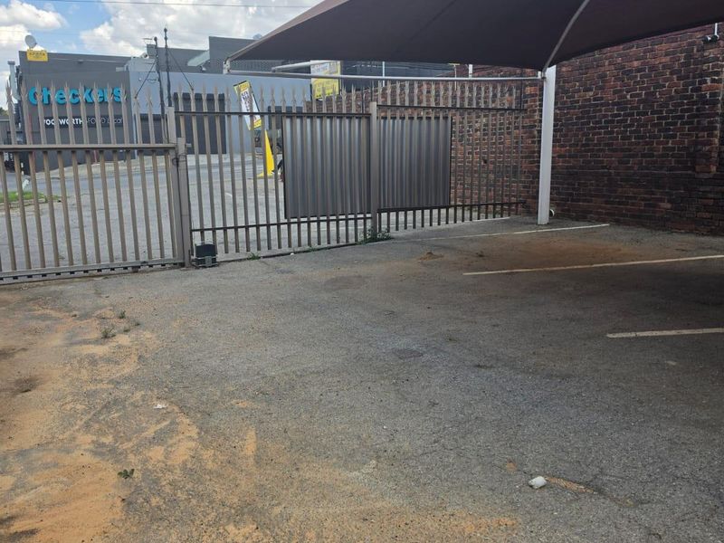 A paved outdoor parking area featuring a shade sail, metal security fencing, and a brick boundary wall.