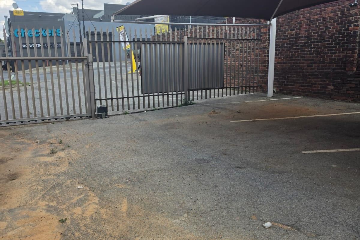 A paved outdoor parking area featuring a shade sail, metal security fencing, and a brick boundary wall.