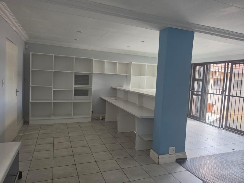 A spacious, empty office area featuring built-in white shelving, a long integrated desk, tiled flooring, and direct access to an outdoor area via sliding doors.