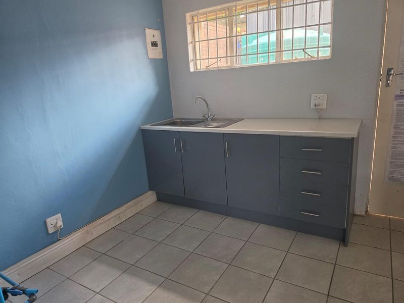 A functional scullery area featuring built-in grey cabinetry, a stainless steel sink, tiled flooring, and a window with security bars.