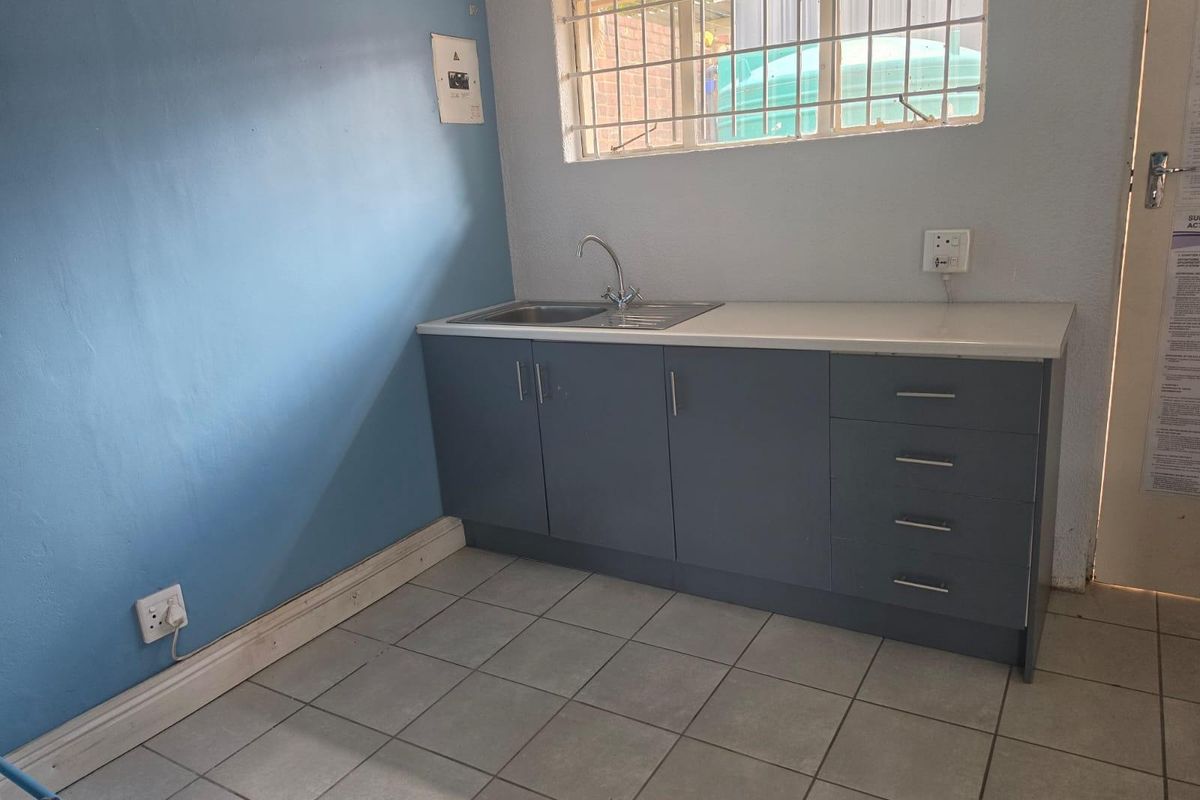 A functional scullery area featuring built-in grey cabinetry, a stainless steel sink, tiled flooring, and a window with security bars.
