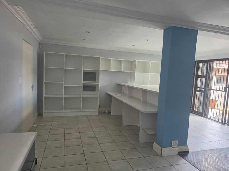 A bright, open-plan office space featuring built-in shelving, a long integrated desk, tiled flooring, and direct access to an outdoor area via a security gate.