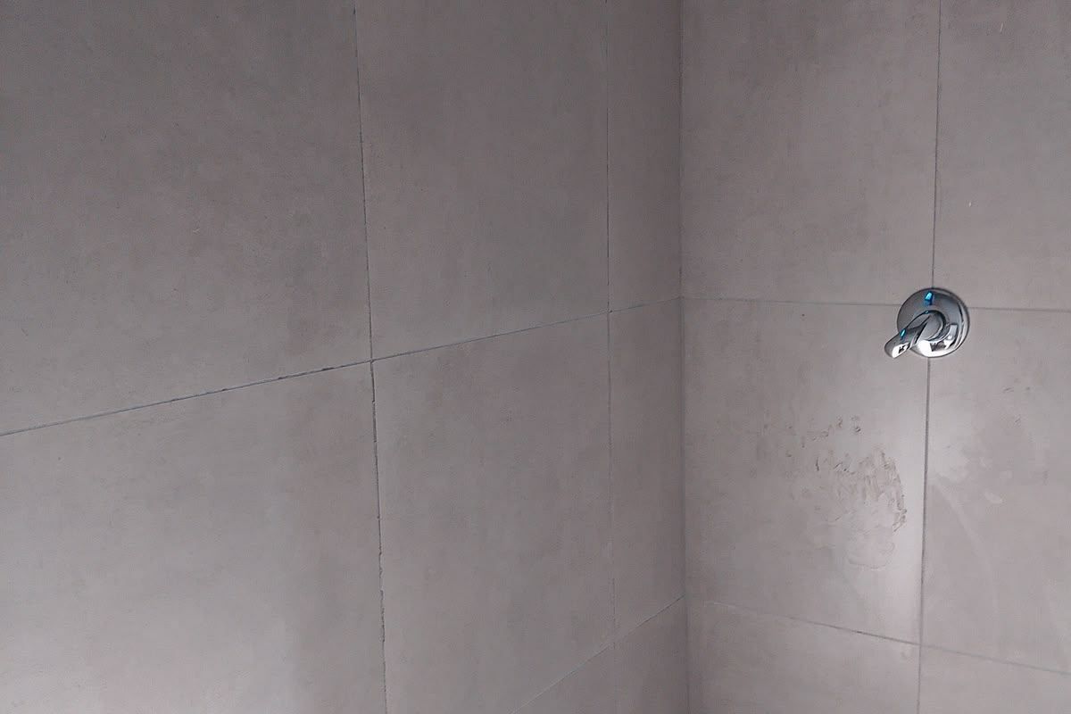 A simple shower enclosure featuring neutral wall and floor tiles, equipped with a standard shower head and mixer tap.
