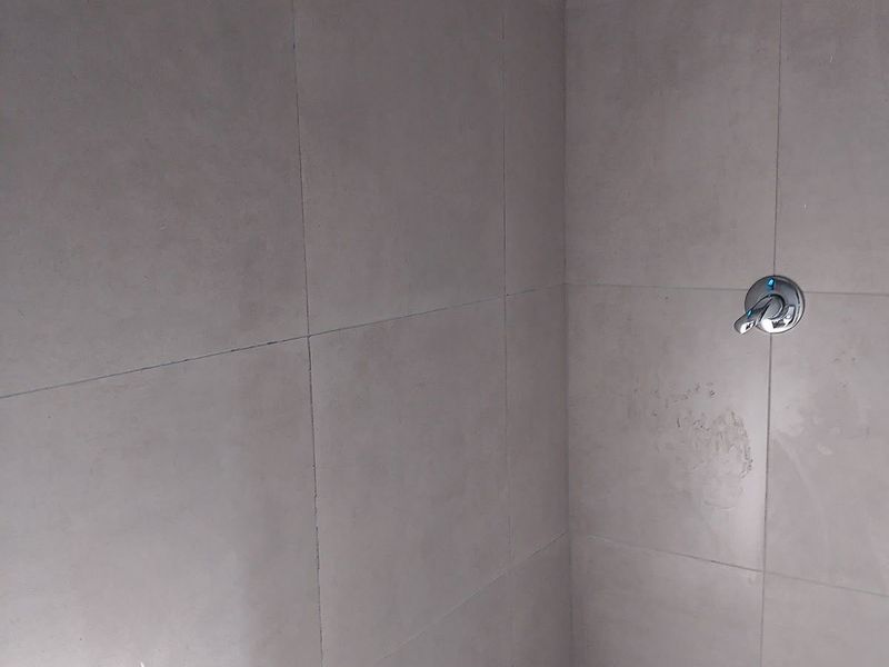 A simple shower enclosure featuring neutral wall and floor tiles, equipped with a standard shower head and mixer tap.