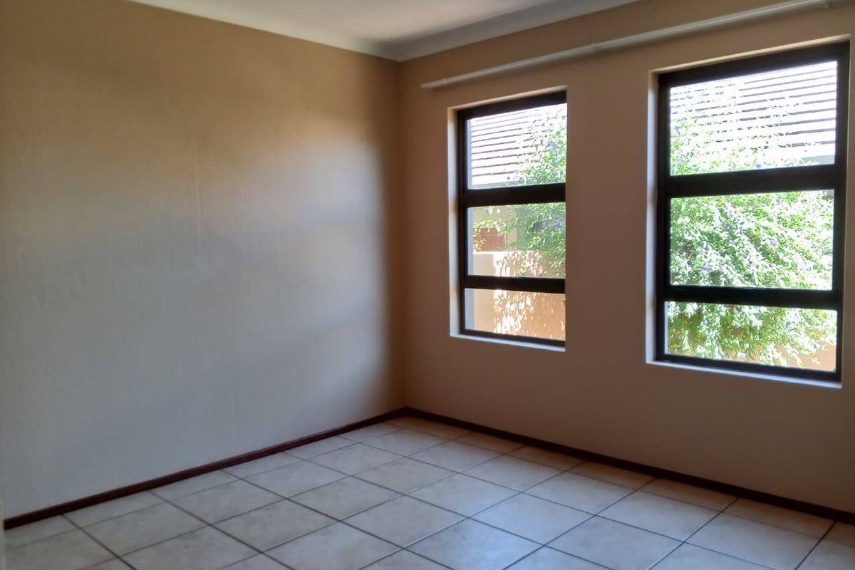 A bright, empty bedroom featuring neutral wall paint, tiled flooring, and large aluminium-framed windows allowing for natural light.