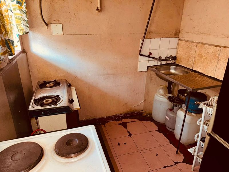 A compact kitchen area featuring a stainless steel sink, tiled splashback, and basic cooking appliances, currently requiring significant renovation and repair to wall and floor finishes.
