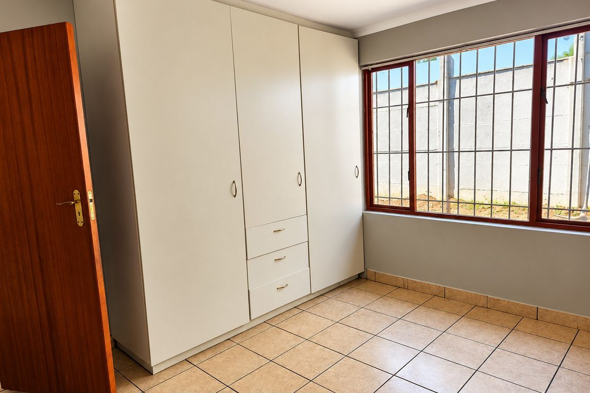 A bright, empty bedroom featuring neutral tiled flooring, built-in cupboards, and a window fitted with security bars.