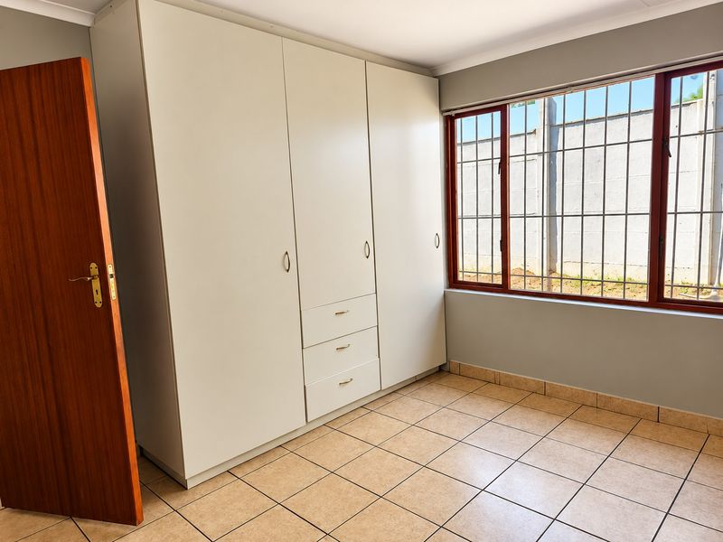A bright, empty bedroom featuring neutral tiled flooring, built-in cupboards, and a window fitted with security bars.