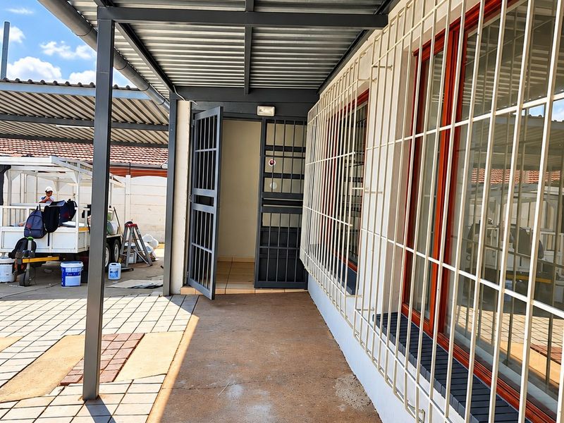 A covered outdoor patio area featuring a corrugated metal roof, security bars on windows, and a secure entrance gate, currently in a functional state.