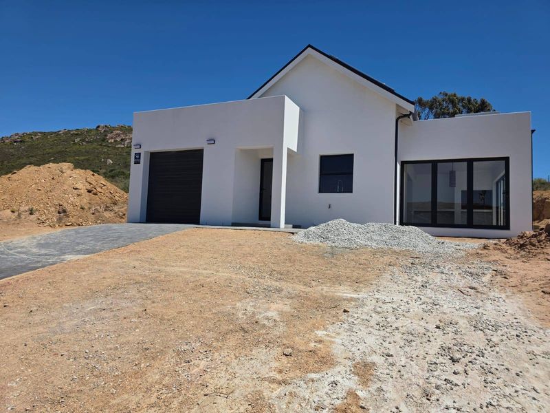 Modern single-story house exterior featuring a garage, paved driveway, and clean white facade, currently undergoing final landscaping and site completion.
