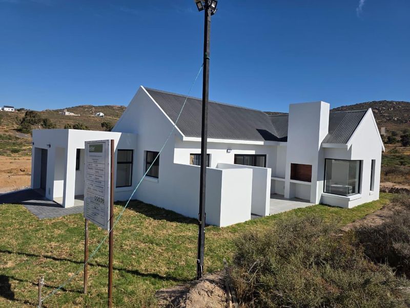 A modern, newly constructed house featuring a clean white facade, corrugated metal roof, and a built-in outdoor braai area on a paved patio.