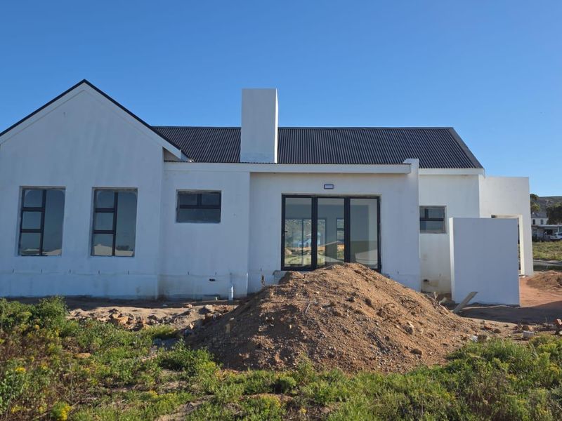 Rear view of a modern single-story house featuring a corrugated metal roof, chimney, and large aluminium-framed windows and doors, currently undergoing final exterior landscaping.
