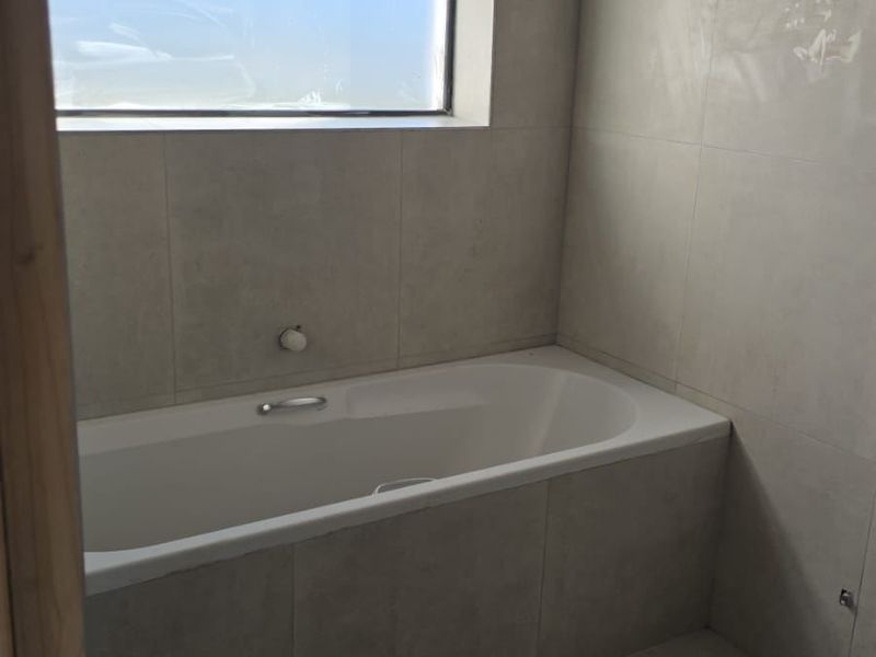 A modern, minimalist bathroom featuring a built-in bathtub, neutral floor-to-ceiling tiling, and a large window providing natural light.