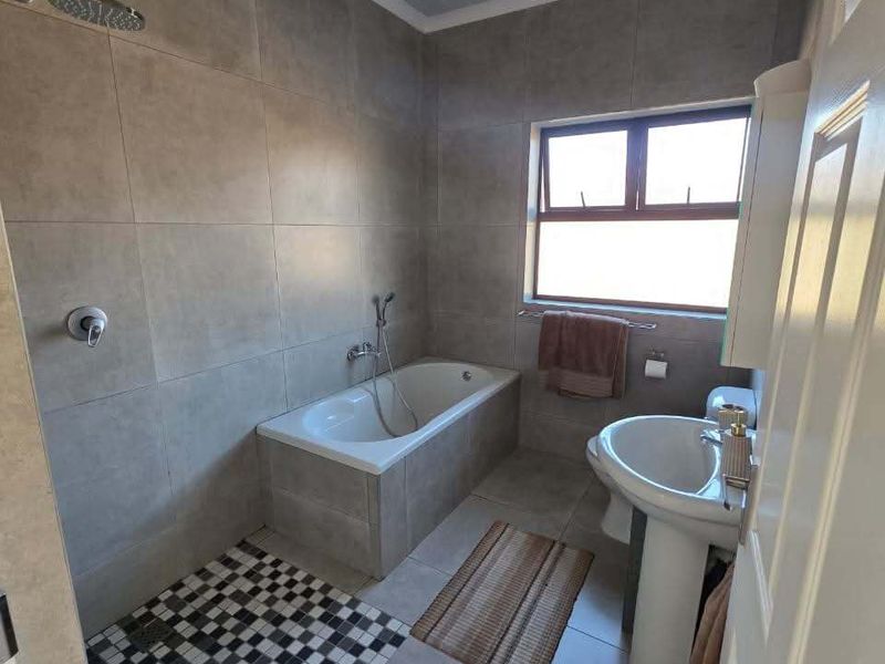 A well-maintained bathroom featuring a built-in bathtub with shower attachment, pedestal sink, toilet, and modern neutral wall tiling with a decorative mosaic floor accent.