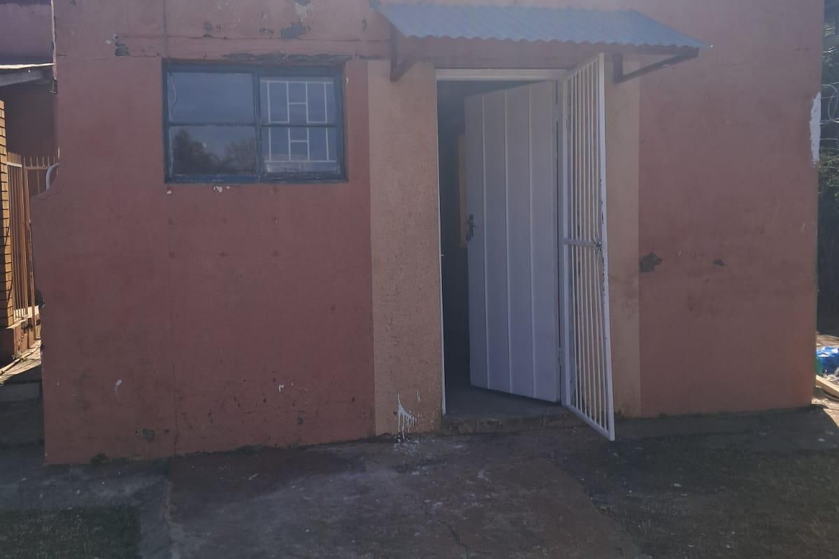 Rear exterior view of the apartment building showing a single entrance door with security gate, small window, and weathered exterior wall finishes.