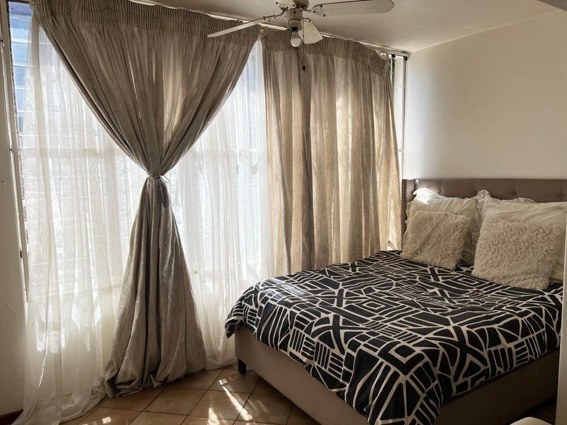 A cozy bedroom featuring tiled flooring, a ceiling fan, and a large window with floor-to-ceiling curtains allowing for natural light.