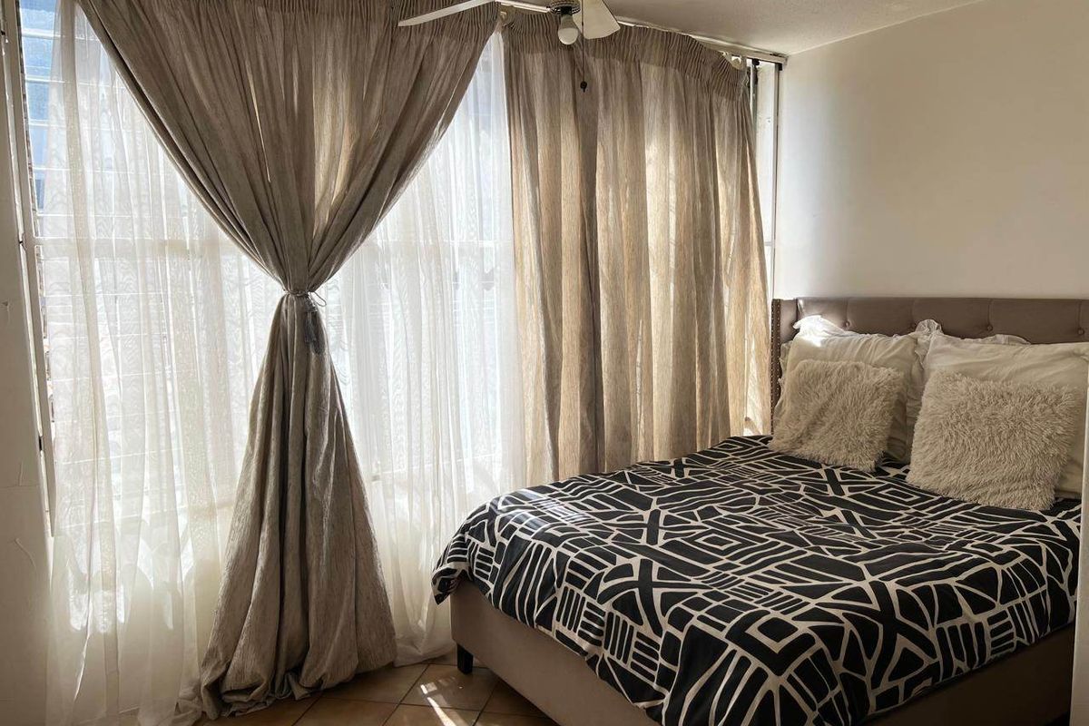 A cozy bedroom featuring tiled flooring, a ceiling fan, and a large window with floor-to-ceiling curtains allowing for natural light.