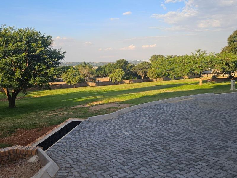A spacious, well-maintained garden area featuring a paved driveway, established trees, and expansive lawn space with a scenic outlook.