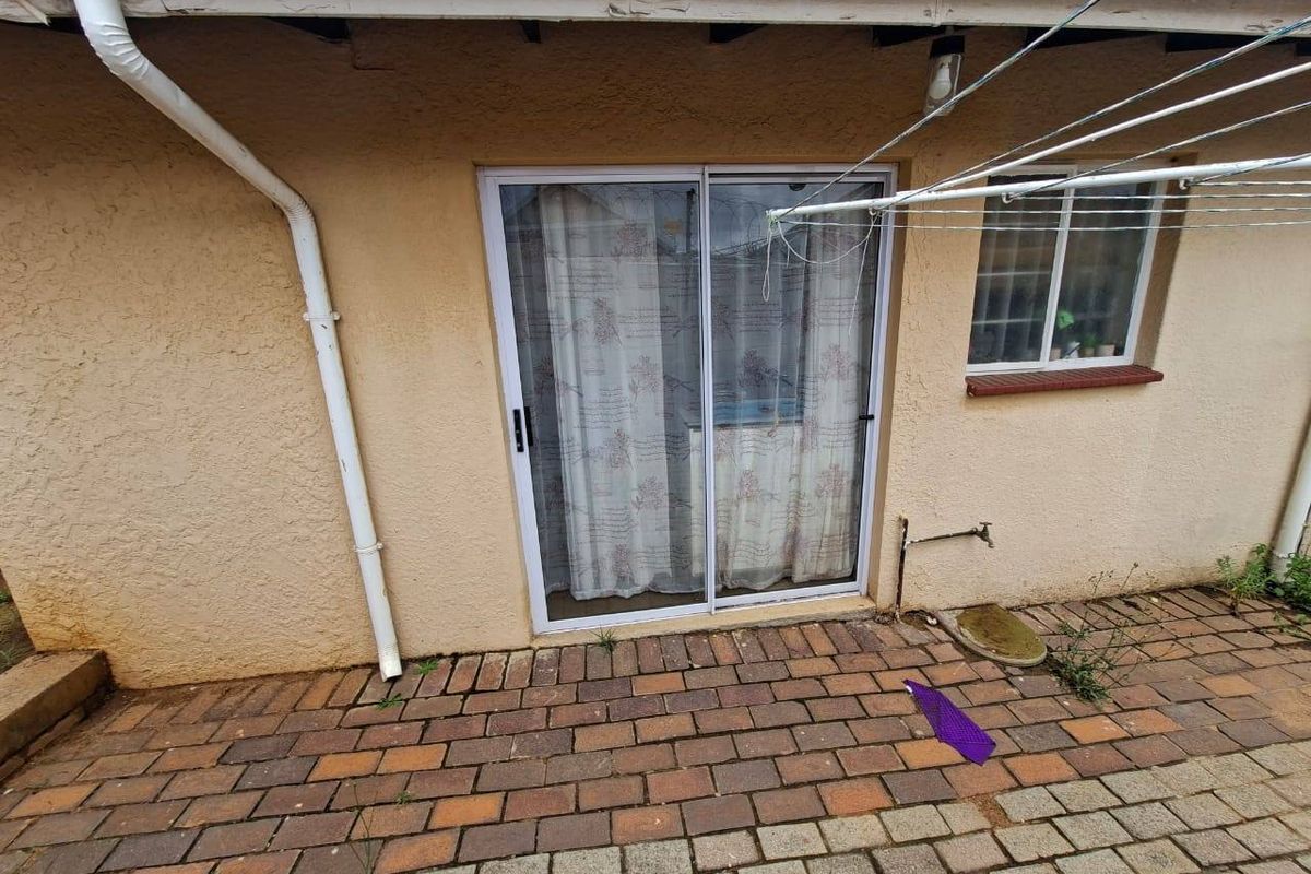 Rear exterior view featuring a paved patio area, sliding glass door access, and external water tap, showing signs of wear on the eaves and walls.