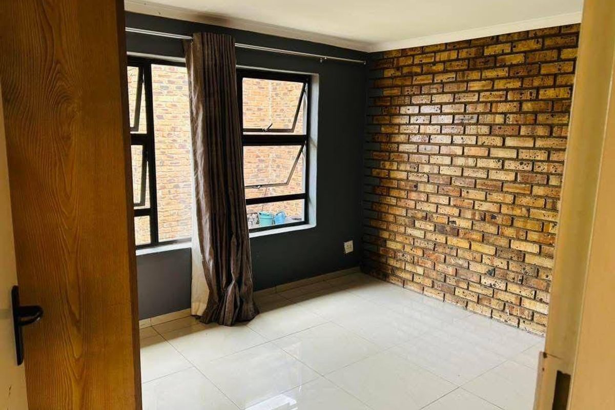 A bright, empty bedroom featuring a rustic exposed brick feature wall, neutral tiled flooring, and modern aluminium-framed windows.