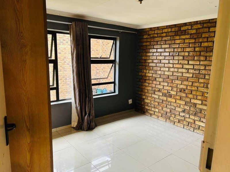 A bright, empty bedroom featuring a rustic exposed brick feature wall, neutral tiled flooring, and modern aluminium-framed windows.